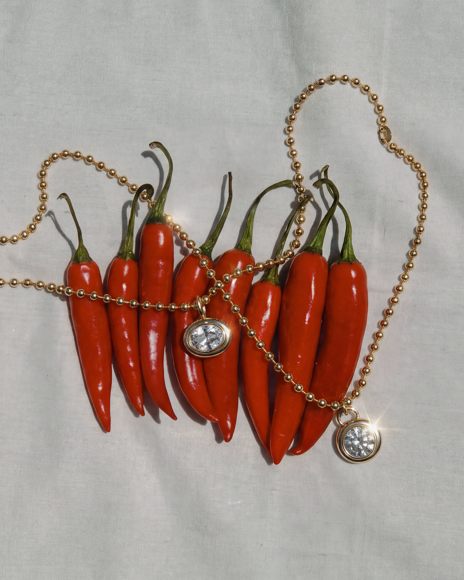 The Mod + Jo Tove Sapphire Bezel Charm, a 14k gold chain necklace with a round sapphire bezel, rests across eight red chili peppers on a light fabric background.