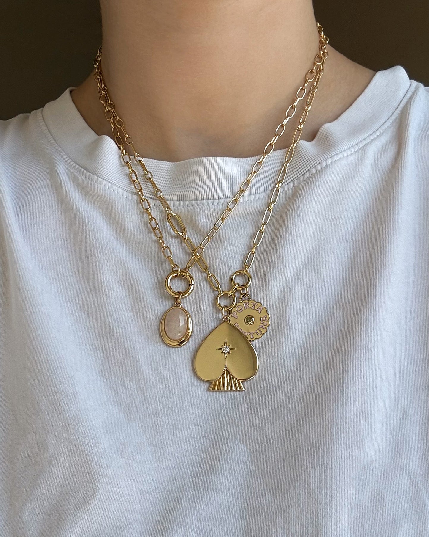 A person wearing a white shirt and layered 14k gold chain necklaces featuring three pendants: a pink oval stone, the Mod + Jo Veya Spade Charm adorned with a small gem, and a round sun charm.