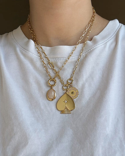 A person wearing a white shirt and layered 14k gold chain necklaces featuring three pendants: a pink oval stone, the Mod + Jo Veya Spade Charm adorned with a small gem, and a round sun charm.