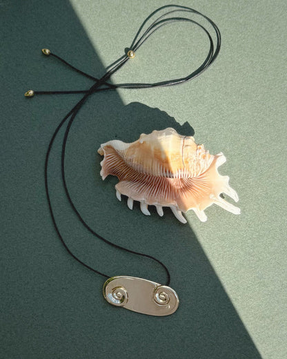 The Suna Spiral Cord Necklace by Mod + Jo, featuring a black adjustable bolo cord and a 14k gold spiral pendant, is displayed with a brown and white seashell on a green surface partially in shadow.