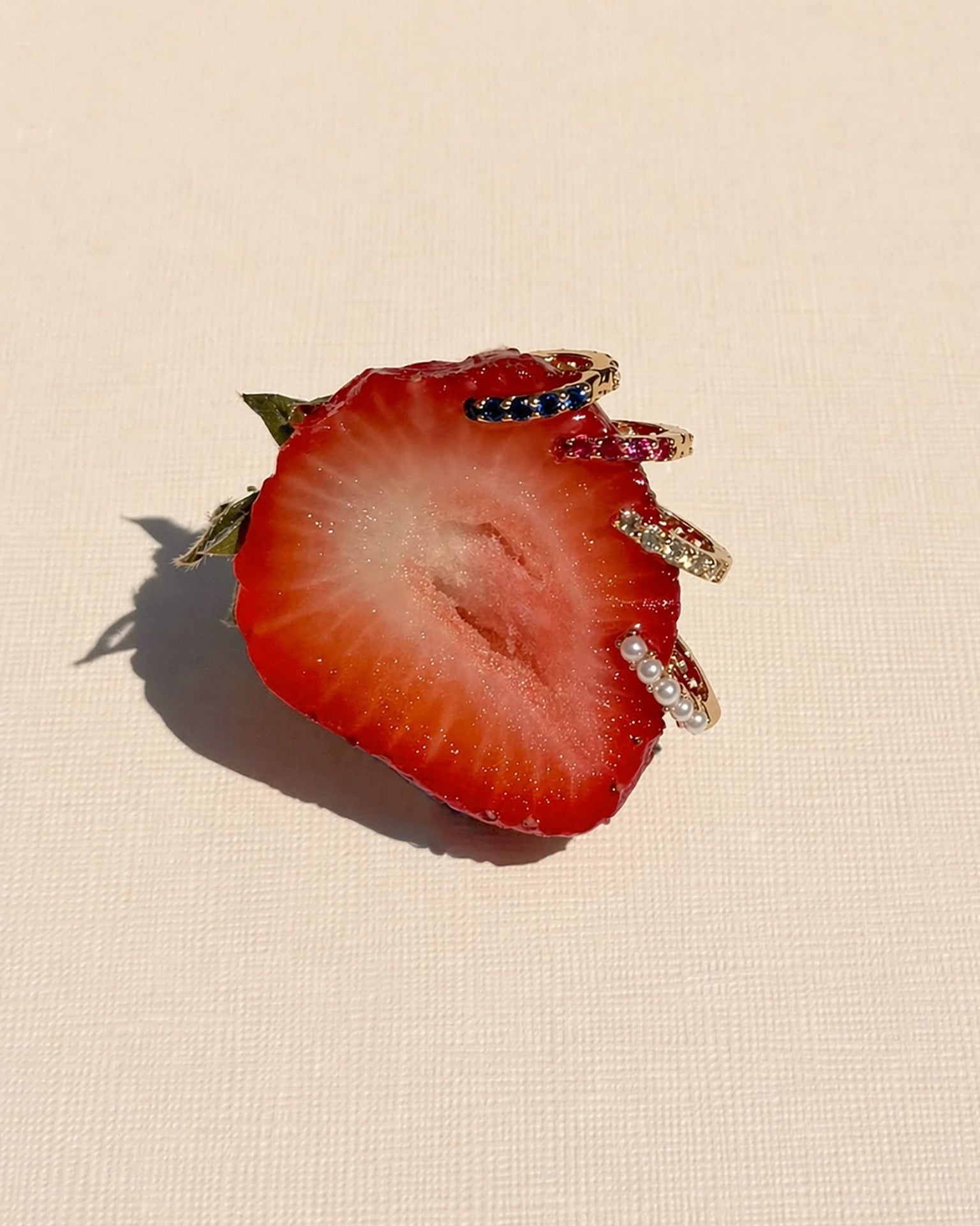 A half strawberry rests on a light textured surface, adorned with the Harlow Reversible Birthstone Huggies by Mod + Jo—gold finish, gem, and pearl embellishments outlining its edge like ornate jewelry.
