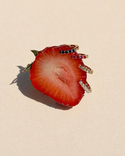 A half strawberry rests on a light textured surface, adorned with the Harlow Reversible Birthstone Huggies by Mod + Jo—gold finish, gem, and pearl embellishments outlining its edge like ornate jewelry.