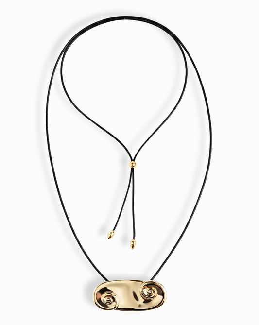 The Suna Spiral Cord Necklace by Mod + Jo features a black cord with an adjustable bolo closure, a sculptural 14k gold pendant with spiral shapes at each end, and two small gold beads on the cord ends.