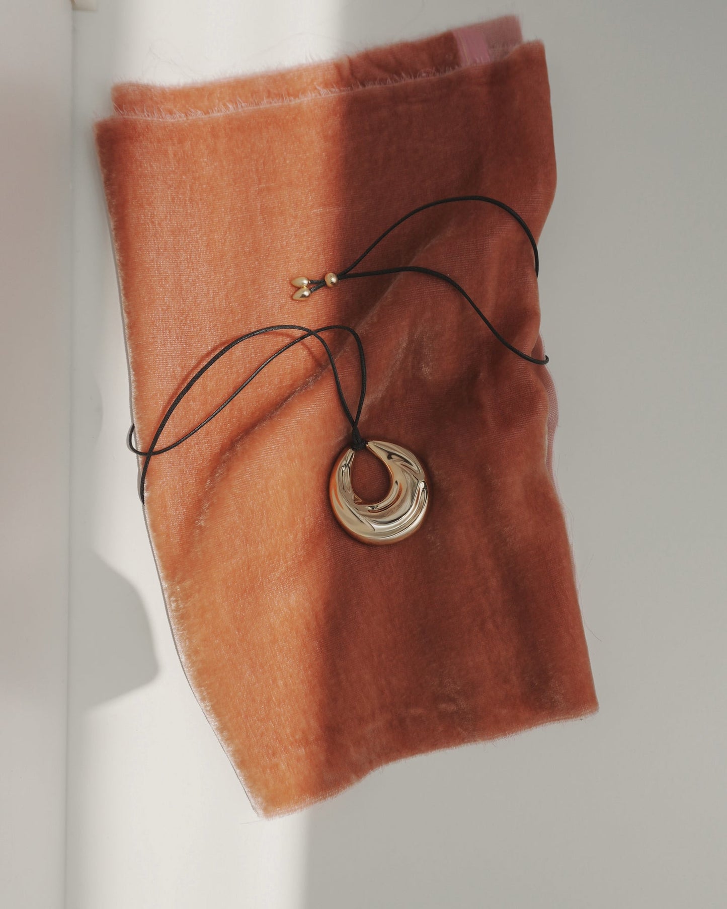 The Kira Cord Necklace by Mod + Jo features a gold pendant on a cord, displayed on soft burnt orange fabric as sunlight creates gentle shadows across the scene.