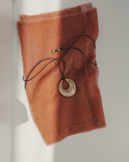 The Kira Cord Necklace by Mod + Jo features a gold pendant on a cord, displayed on soft burnt orange fabric as sunlight creates gentle shadows across the scene.