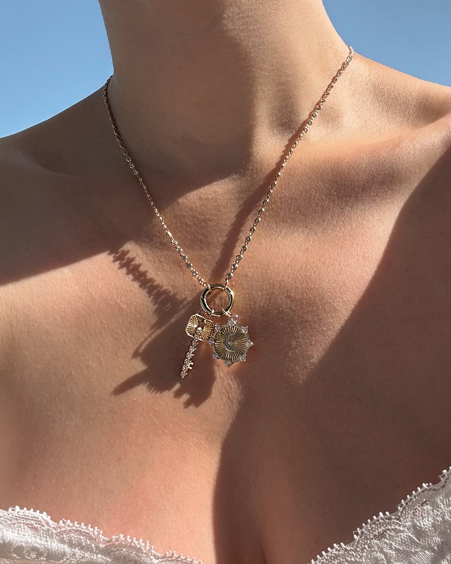 A close-up of a person’s neck wearing Mod + Jo’s Noah Key Charm—a gold plated necklace adorned with key and flower-shaped charms—against a clear blue sky and dressed in white lace, showcasing an elegant piece of symbolic jewelry that beautifully blends style and meaning.