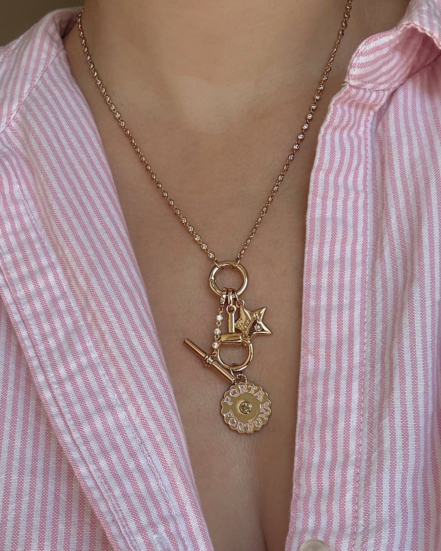 A person wearing a pink and white striped shirt and a gold necklace adorned with charms, including a key, padlock, the Mod + Jo Elara Lucky Star Charm, and a round Louis Vuitton pendant. The necklace showcases elegant 14k gold jewelry details for a chic finishing touch.