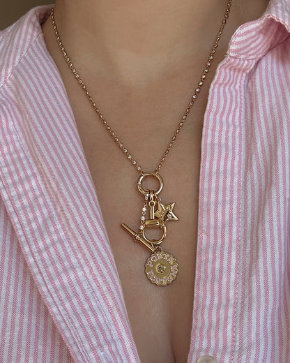 A person wearing a pink and white striped shirt and a gold necklace adorned with charms, including a key, padlock, the Mod + Jo Elara Lucky Star Charm, and a round Louis Vuitton pendant. The necklace showcases elegant 14k gold jewelry details for a chic finishing touch.