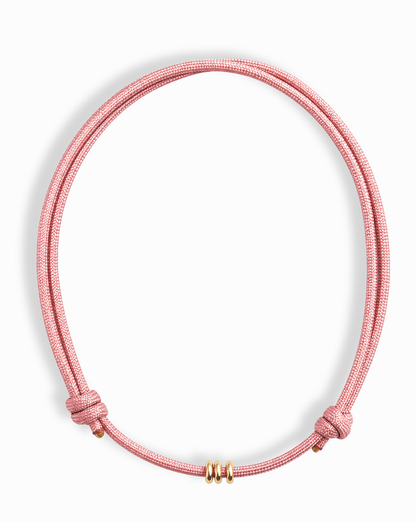 The Tommy Cord Necklace by Mod + Jo features a pink paracord with three gold-plated spacers at the front and adjustable side knots, shown against a white background.
