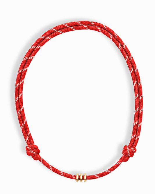 The Mod + Jo Tommy Cord Necklace features a simple red cord with white diagonal stripes, knotted at each end and adorned with three small gold rings at the center, shown against a white background.