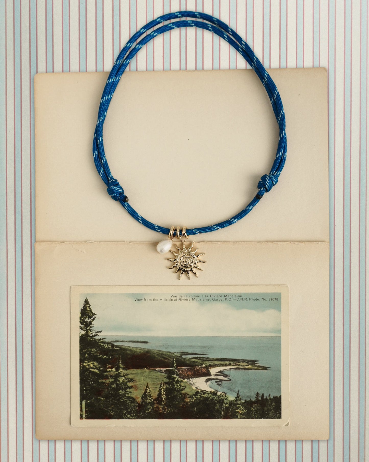The Tommy Cord Necklace by Mod + Jo, featuring a sun-shaped pendant, pearl, and gold-plated spacer on blue paracord, is displayed atop an open envelope with a vintage coastal postcard and striped pastel paper in the background.