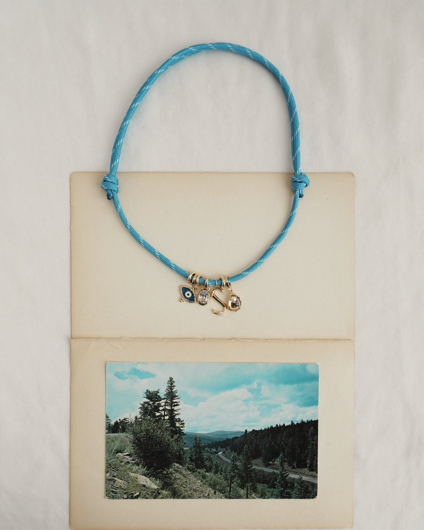 The Mod + Jo Tommy Cord Necklace, featuring gold-plated spacers and charms, is displayed on an open vintage postcard with a scenic landscape of trees and hills against a partly cloudy sky. The background is simple white fabric.