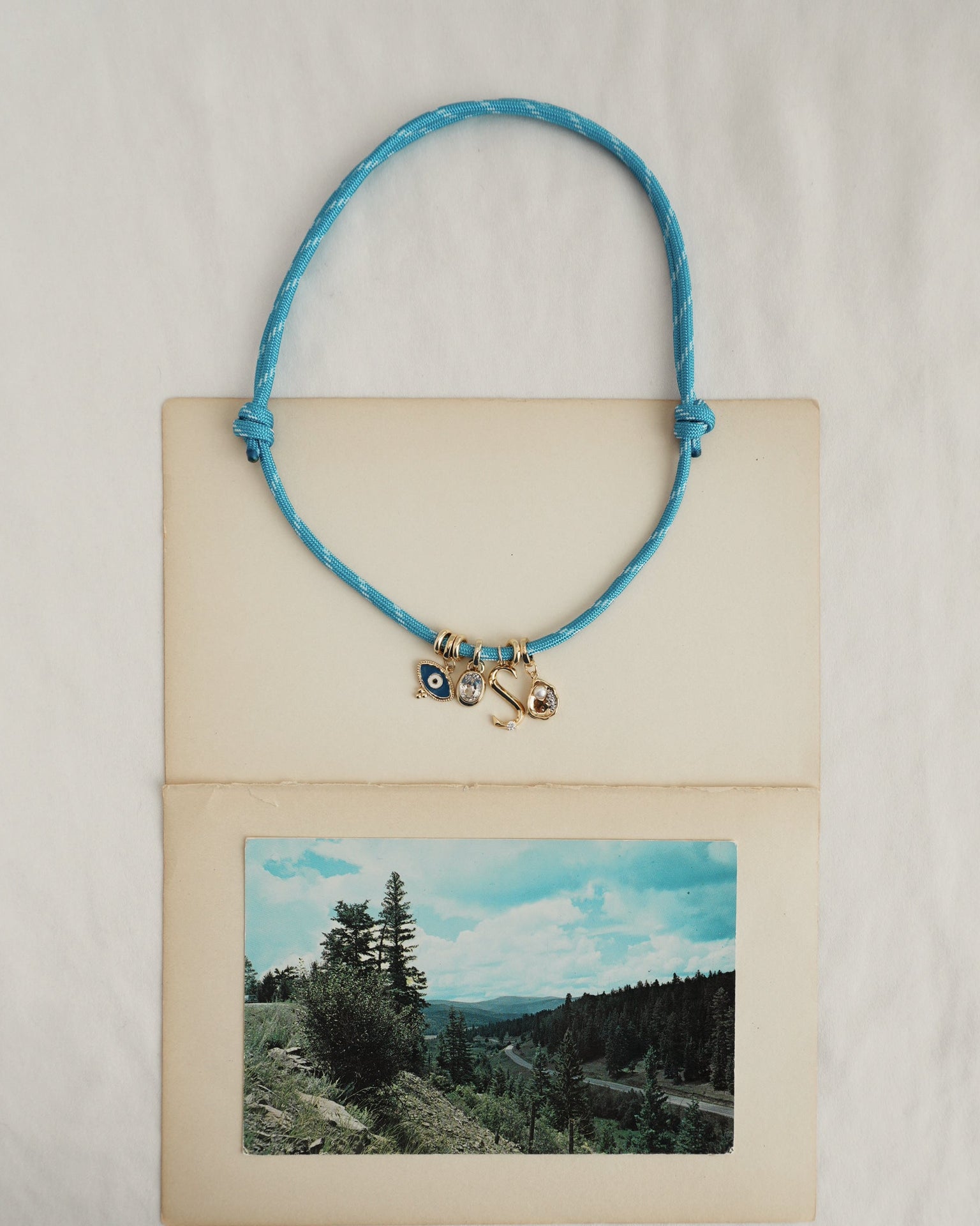 The Mod + Jo Tommy Cord Necklace, featuring gold-plated spacers and charms, is displayed on an open vintage postcard with a scenic landscape of trees and hills against a partly cloudy sky. The background is simple white fabric.