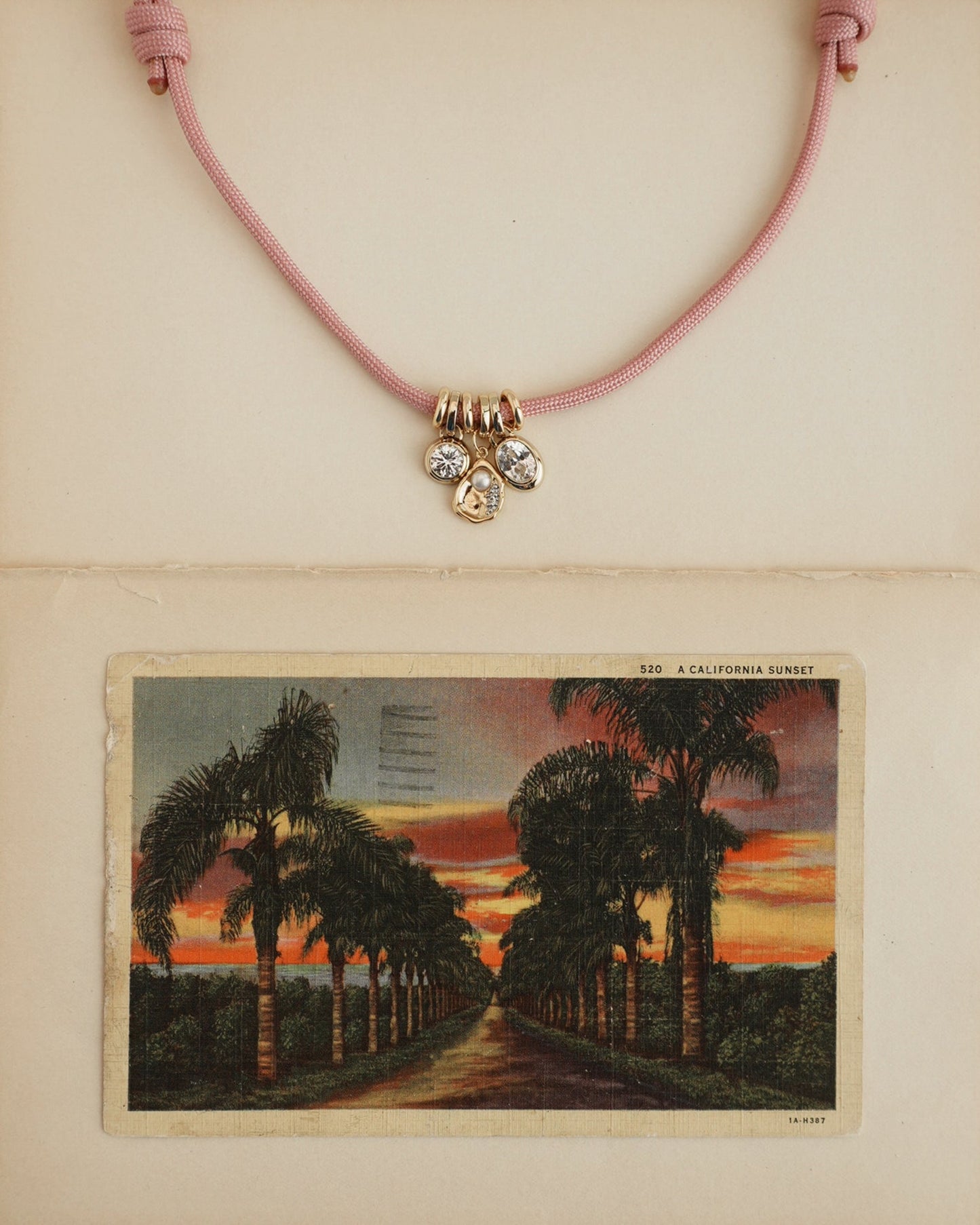 The Tommy Cord Necklace by Mod + Jo, featuring pink paracord, three gemstone charms, and gold-plated spacers, is displayed above a vintage postcard of a palm-lined sunset road on a cream-colored background.