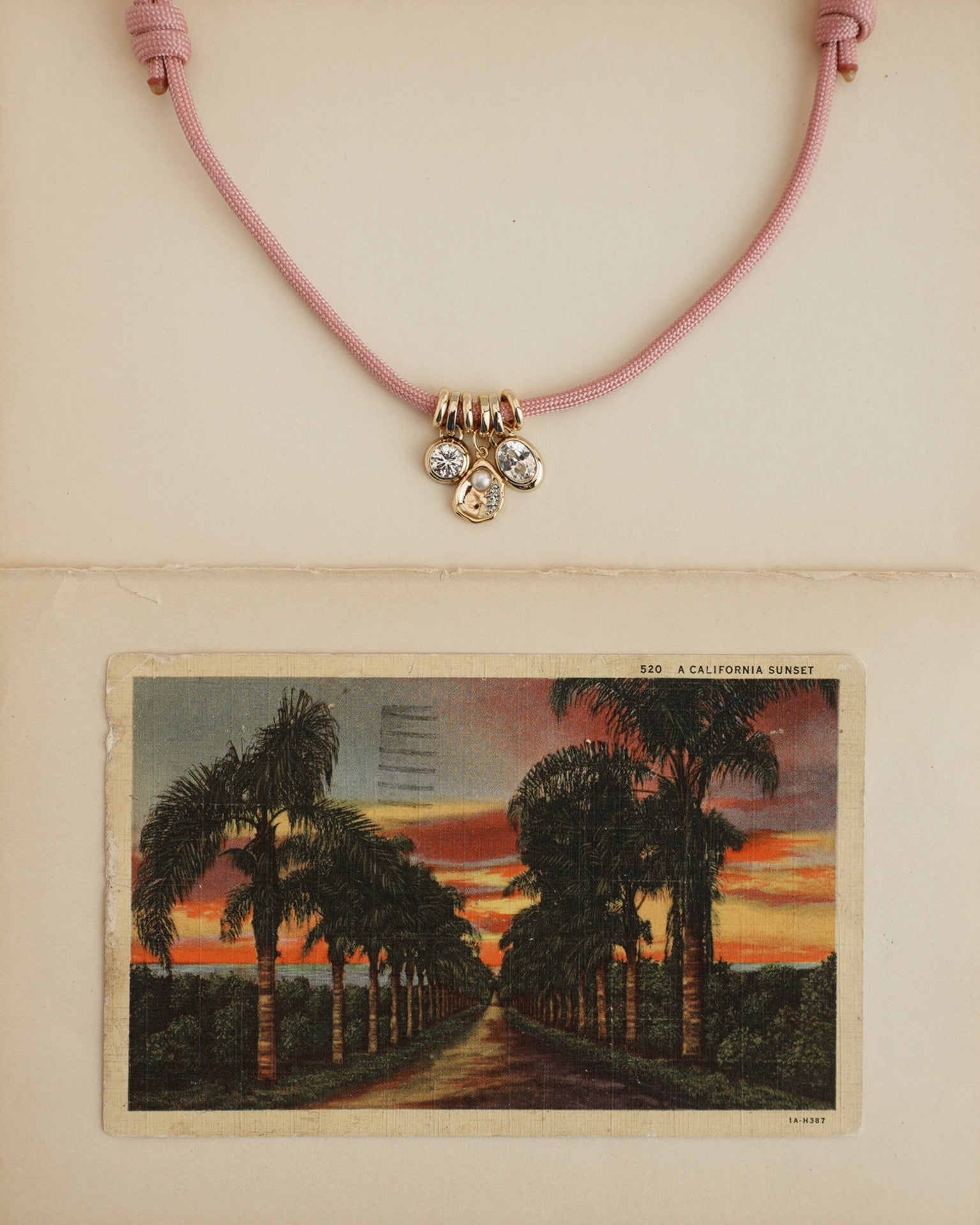 The Tommy Cord Necklace by Mod + Jo, featuring pink paracord, three gemstone charms, and gold-plated spacers, is displayed above a vintage postcard of a palm-lined sunset road on a cream-colored background.