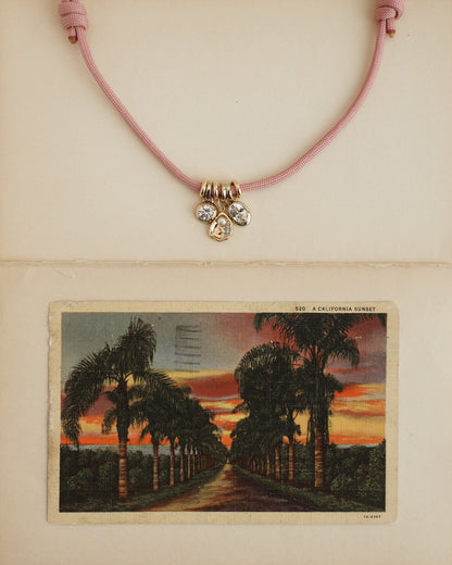 The Tommy Cord Necklace by Mod + Jo, featuring pink paracord, three gemstone charms, and gold-plated spacers, is displayed above a vintage postcard of a palm-lined sunset road on a cream-colored background.