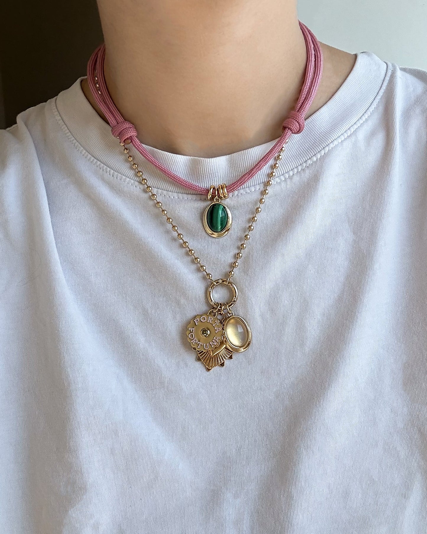 A person wearing a white shirt layered with two necklaces: a pink cord featuring Mod + Jo’s Rory Gemstone Charm—a green oval pendant that shimmers like moonstone—and a gold chain adorned with round gold charms and a small pearl-like locket.