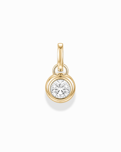 The Tove Sapphire Bezel Charm by Mod + Jo is a small 14k gold pendant featuring a round, clear lab-grown sapphire in a simple bezel setting with a gold loop, shown on a white background.