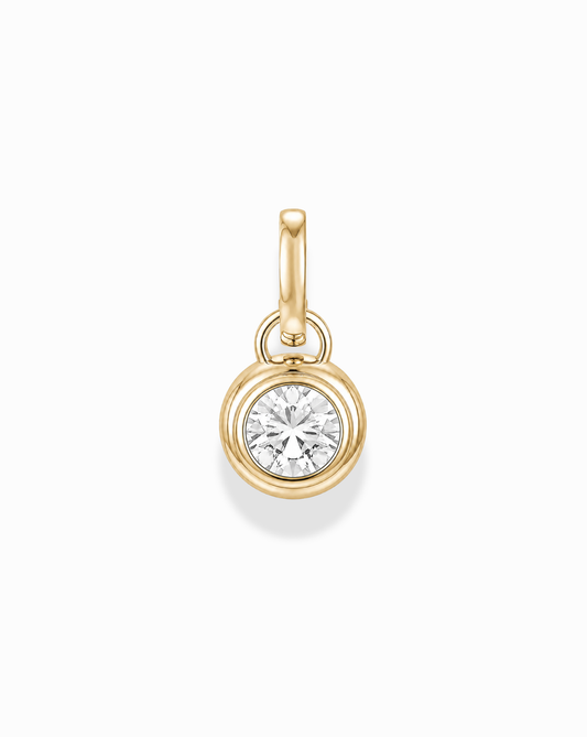 The Tove Sapphire Bezel Charm by Mod + Jo is a small 14k gold pendant featuring a round, clear lab-grown sapphire in a simple bezel setting with a gold loop, shown on a white background.