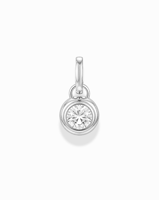 The Mod + Jo Tove Sapphire Bezel Charm is a silver pendant with a round, bezel-set lab-grown white sapphire and a small loop for attaching to necklaces or bracelets, shown on a white background.