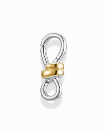 The Izzy Knot Charm Enhancer by Mod + Jo features a silver metal figure-eight clasp, highlighted by two intertwined 14k gold-plated loops at the center, displayed on a white background.