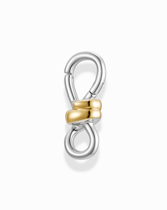 The Izzy Knot Charm Enhancer by Mod + Jo features a silver metal figure-eight clasp, highlighted by two intertwined 14k gold-plated loops at the center, displayed on a white background.