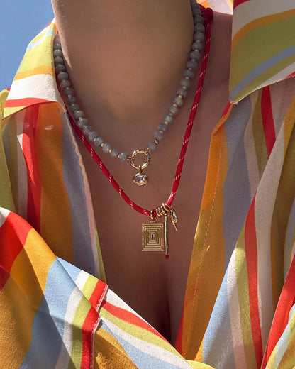 A person wears a vibrant striped shirt, a beaded necklace with a round gold pendant, and the Mod + Jo Tommy Cord Necklace—an adjustable paracord style with gold geometric pendants. The close-up highlights both the jewelry and colorful shirt.