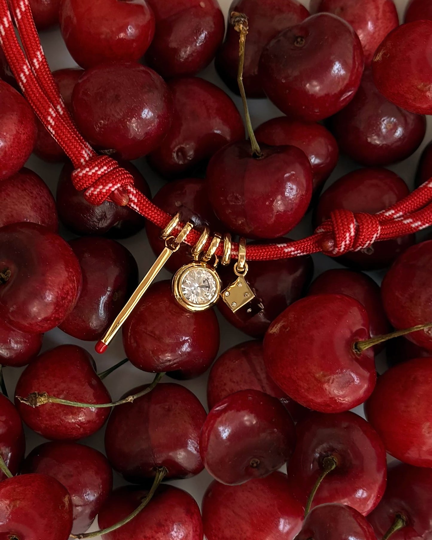 A close-up of fresh red cherries with a Tommy Cord Necklace by Mod + Jo—a red string piece adorned with playful gold charms like a matchstick, round gem, and dice—showcasing its adjustable, whimsical design.
