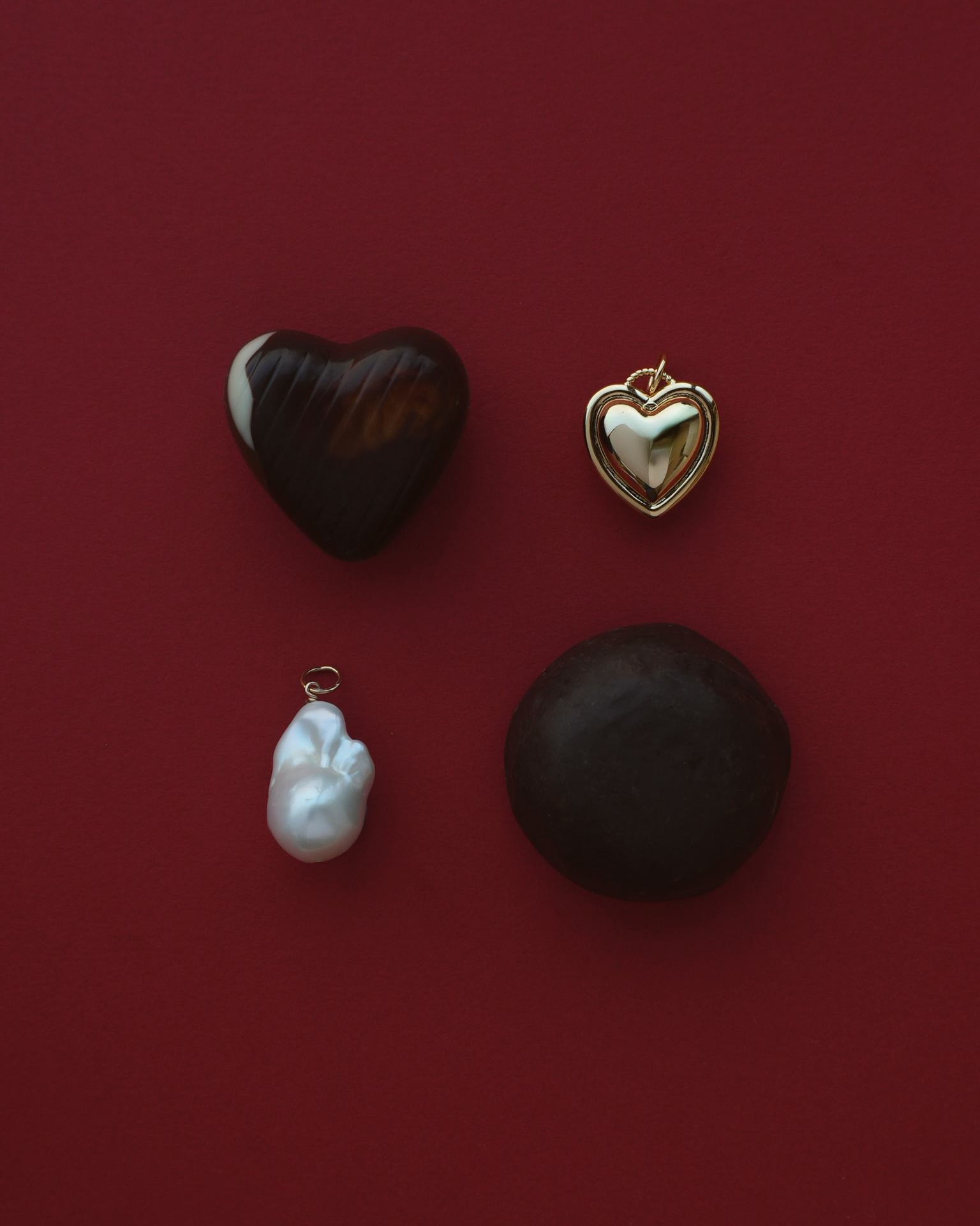 A gold heart charm and a large baroque pearl charm laying on a red background with two valentine's day style chocolates