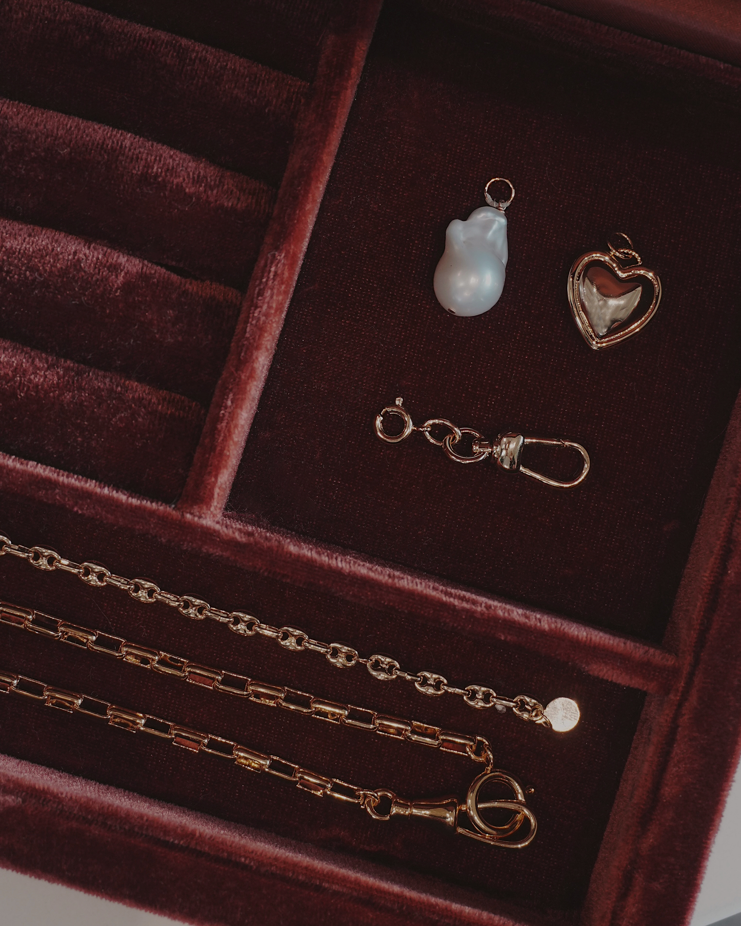 Jewelry laying in a jewelry box including a three gold chain, a heart pendant, a pearl charm. and a charm clip, all laying on a dark red velvet surface.