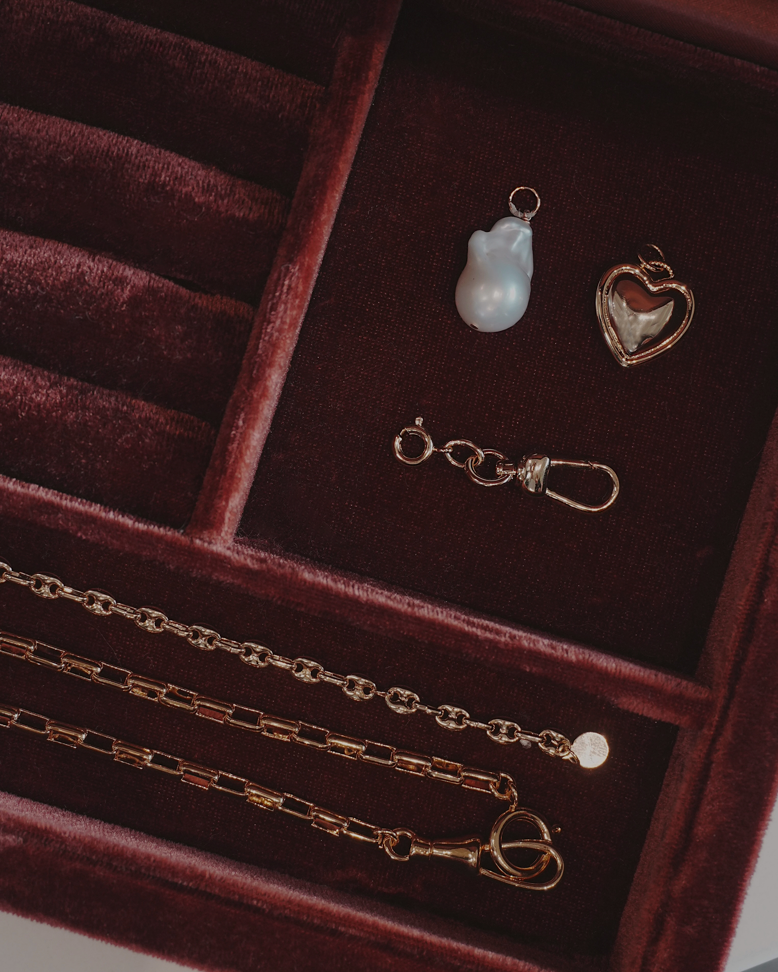 Jewelry laying in a jewelry box including a three gold chain, a heart pendant, a pearl charm. and a charm clip, all laying on a dark red velvet surface.