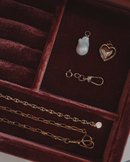 Jewelry laying in a jewelry box including a three gold chain, a heart pendant, a pearl charm. and a charm clip, all laying on a dark red velvet surface.